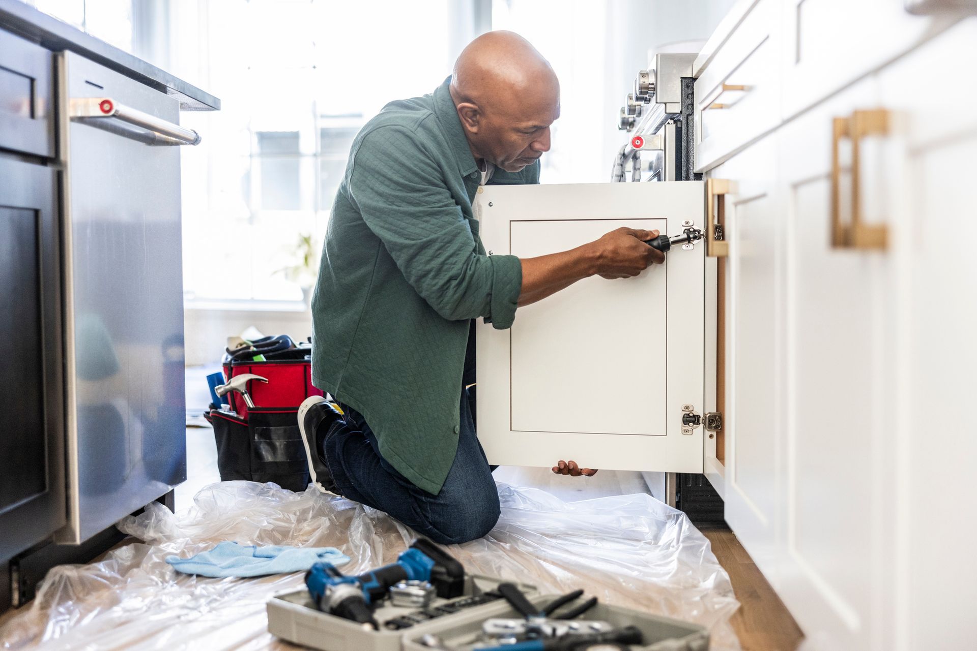 A man is kneeling on the floor fixing a cabinet in a kitchen.