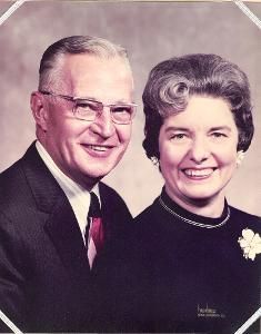 Couple smiling, posed. Man in suit, glasses. Woman in dark dress, pearls, floral pin.