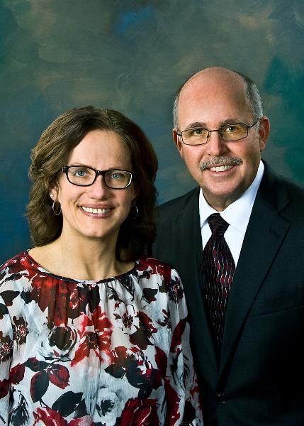 A smiling couple poses for a portrait. The woman wears glasses and a floral shirt. The man wears a suit and tie.