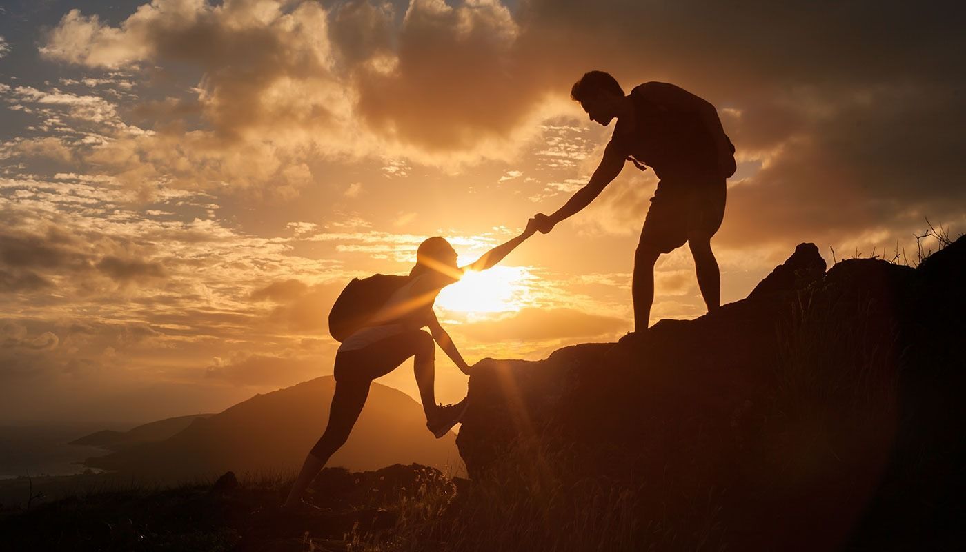 A man is helping another man climb a mountain at sunset.