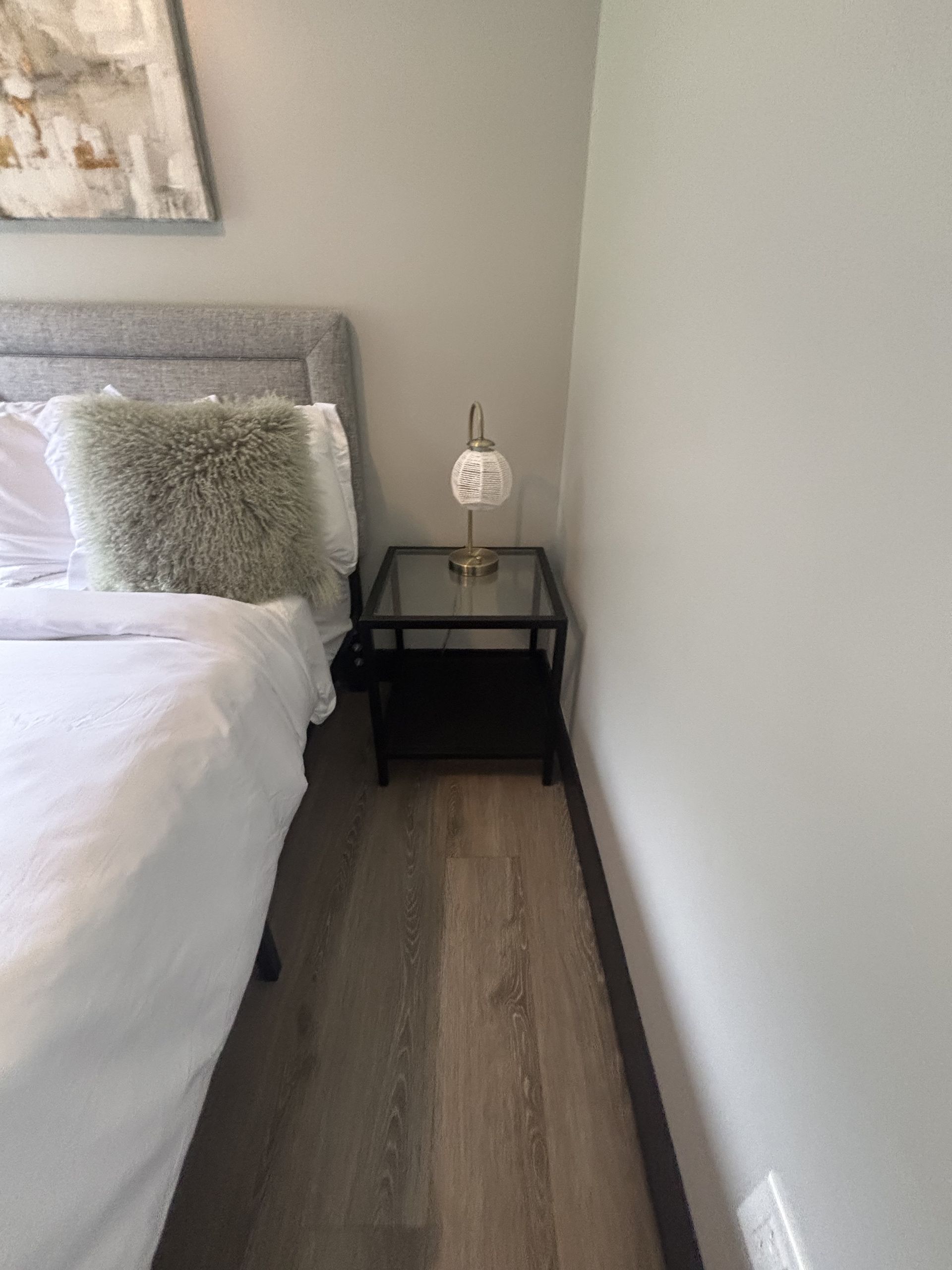 Bedside table with lamp next to a bed with white linens and a fluffy pillow.