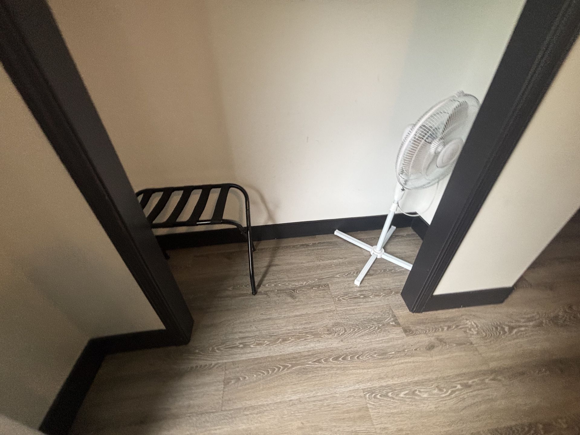 A small space with a folding luggage rack and a white fan; walls are white with dark trim.