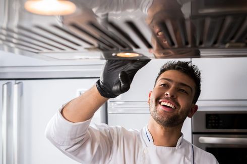 Chef smiling while cleaning a stainless steel kitchen hood with a gloved hand