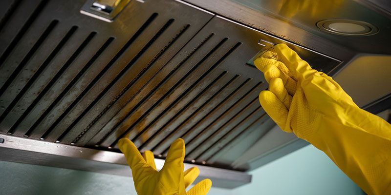 Gloved hands cleaning a stainless steel kitchen hood filter underneath