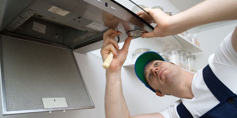 Technician in hard hat inspecting a ceiling vent with a flashlight and tool inside a utility room