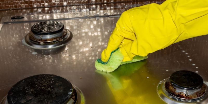 Yellow-gloved hand wiping a stainless steel stovetop with a green sponge