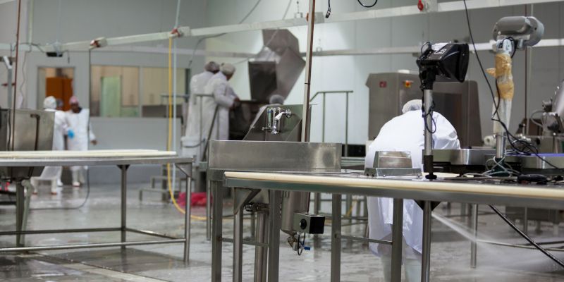 Workers in white lab coats in a stainless-steel industrial cleanroom, handling equipment with machinery overhead