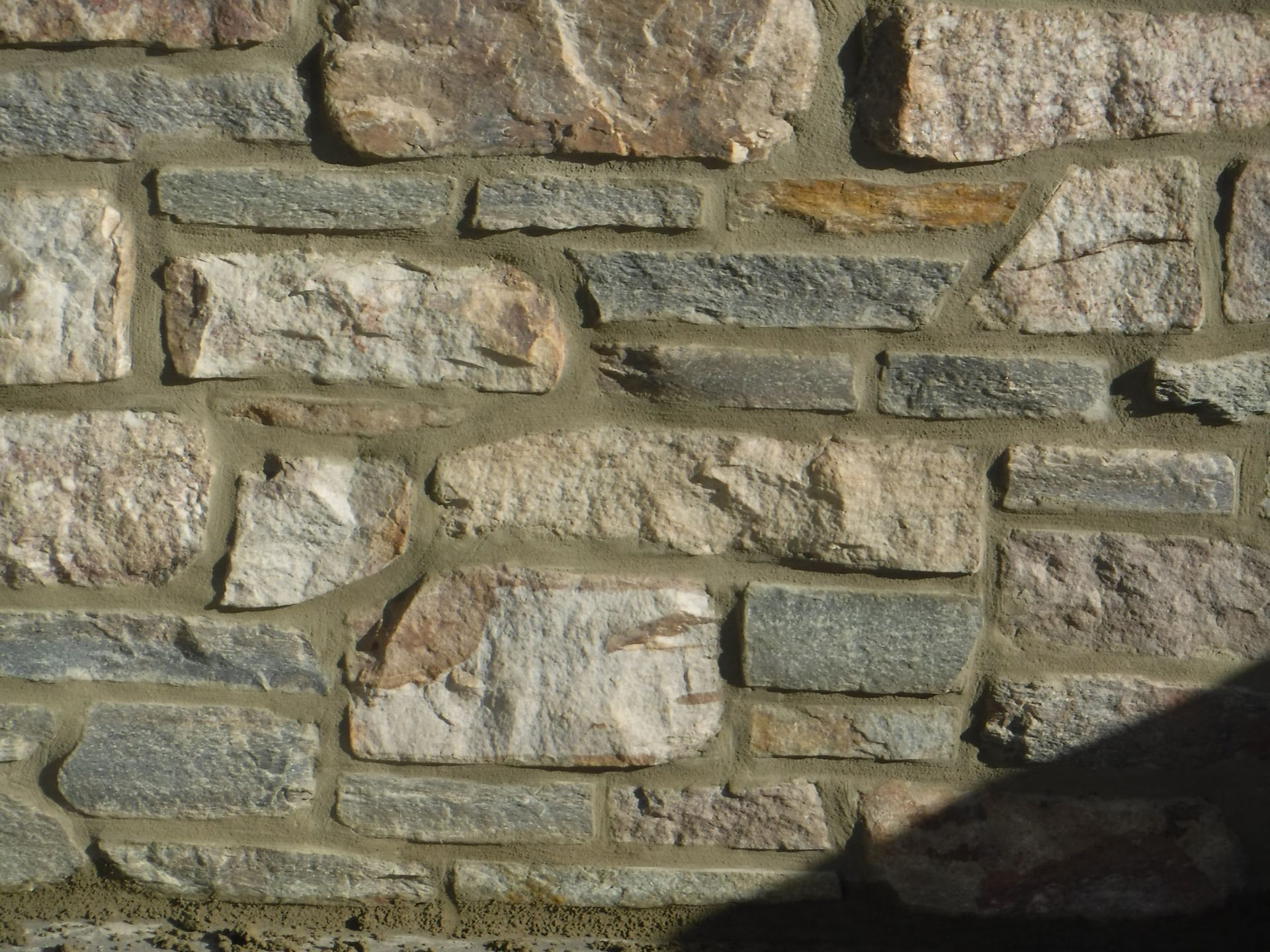 Stone wall made of irregular tan and gray rocks with mortar lines and a dark shadow in the lower right