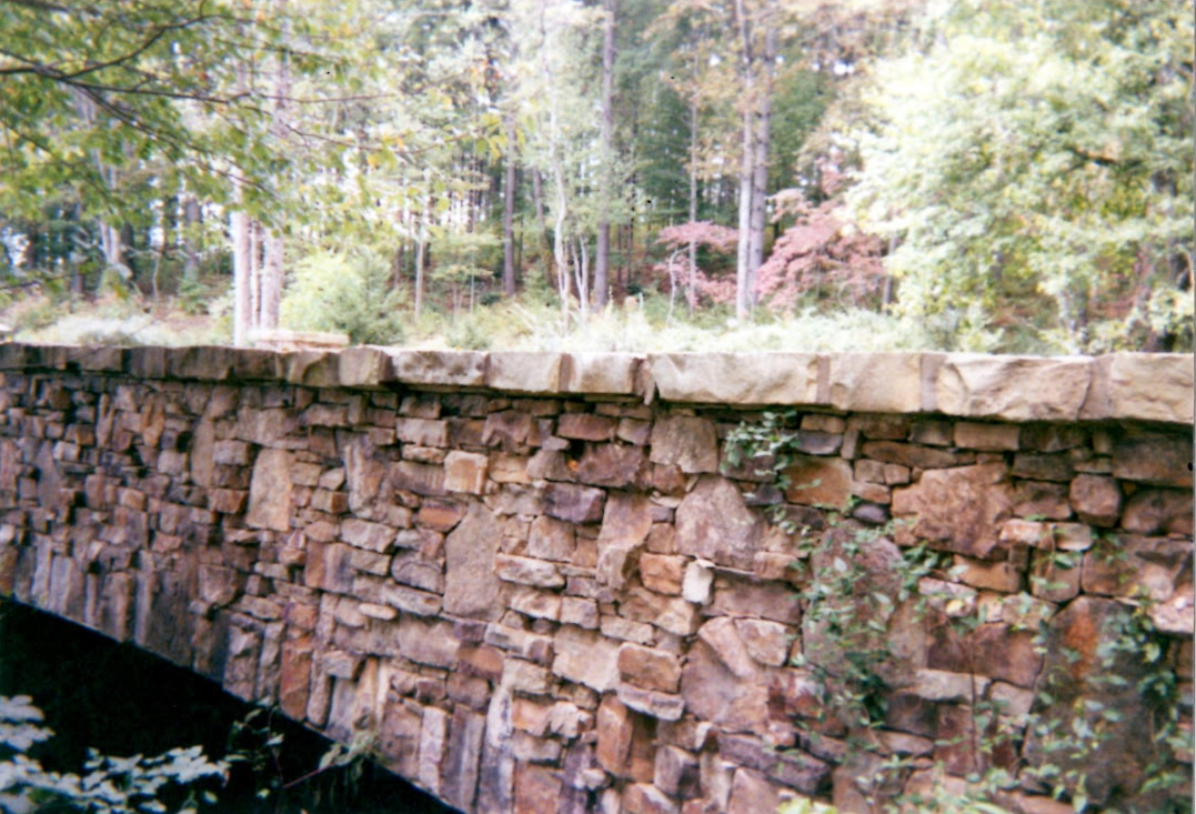 Stone bridge wall over water in a wooded park, with trees and greenery above.