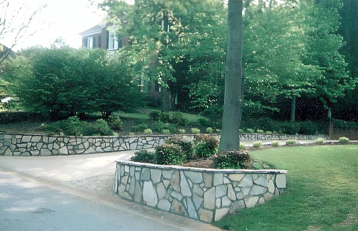 Stone planter with a tree in a landscaped garden beside a paved walkway, surrounded by greenery