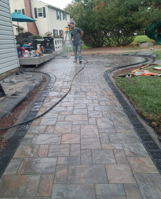 Worker pressure washing a curved brick walkway beside a house and garden