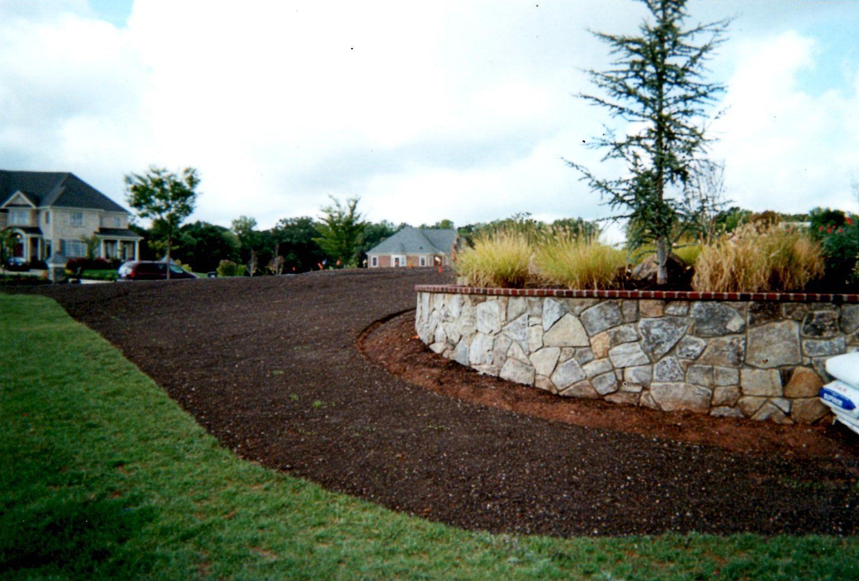 Landscaped yard with stone retaining wall, brown mulch path, and houses in the background