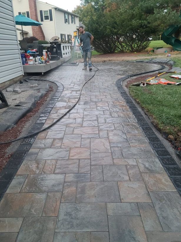 Stone walkway leading past a house and garden, with a person standing near a bench on the left.