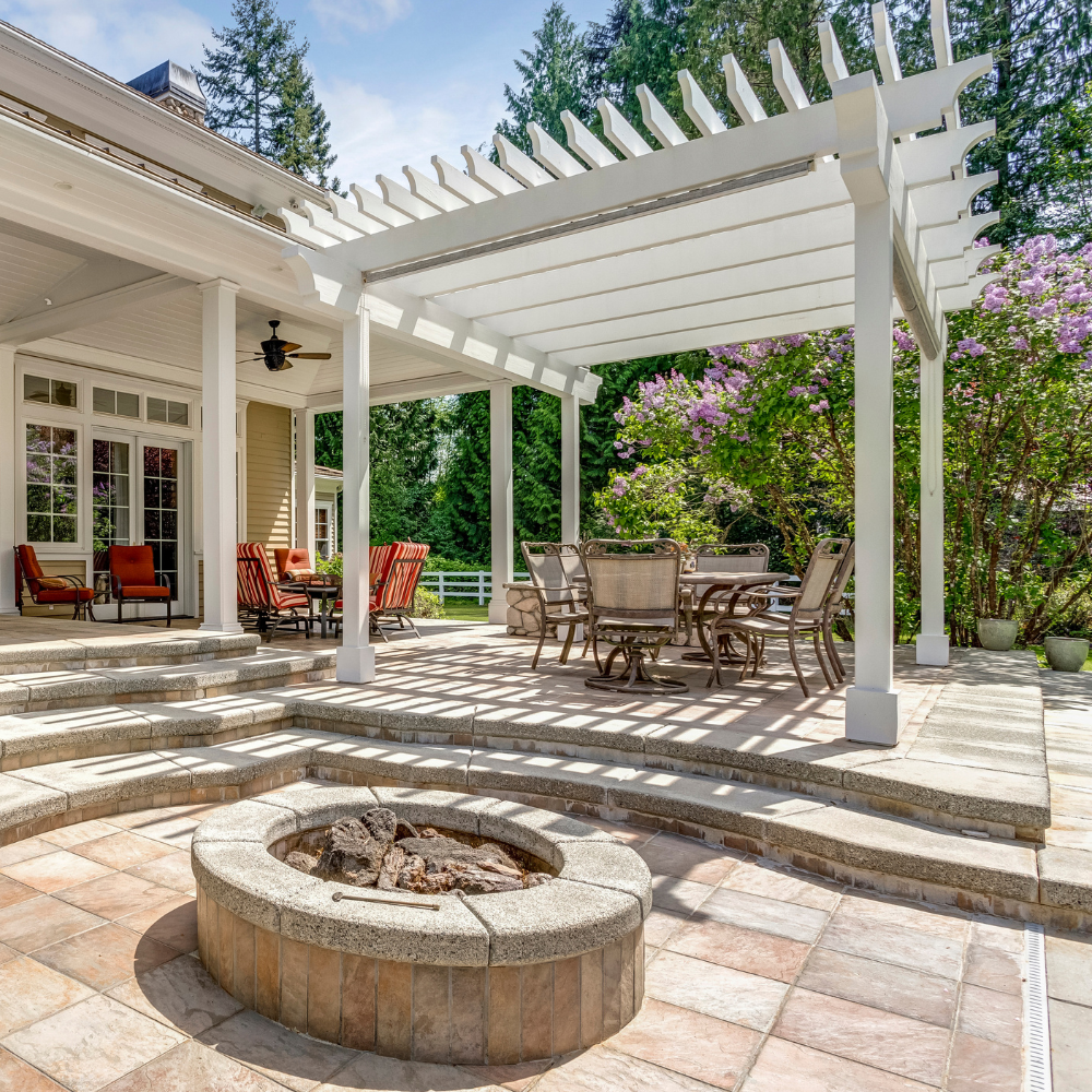 Sunlit backyard patio with white pergola, stone fire pit, outdoor dining set, and house exterior
