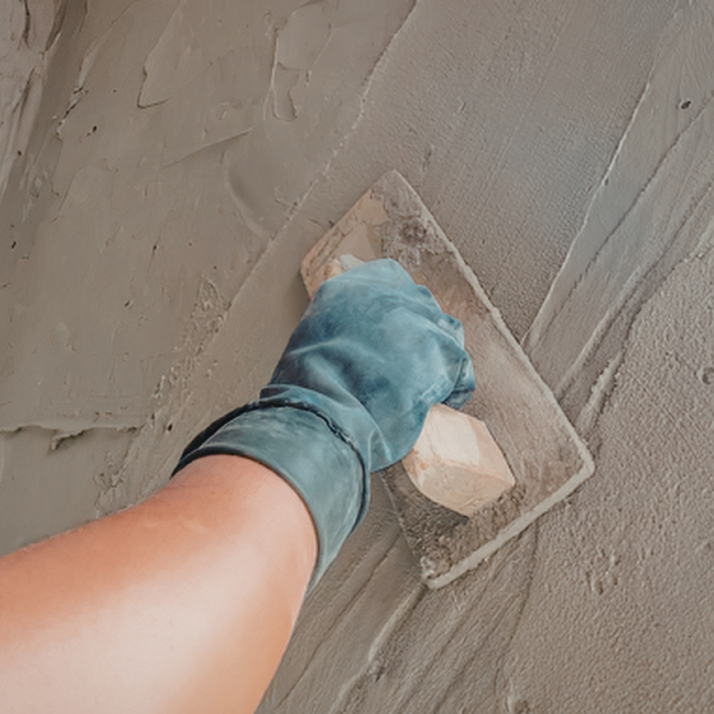 Hand trowel smoothing gray plaster on a wall, with a gloved hand in view