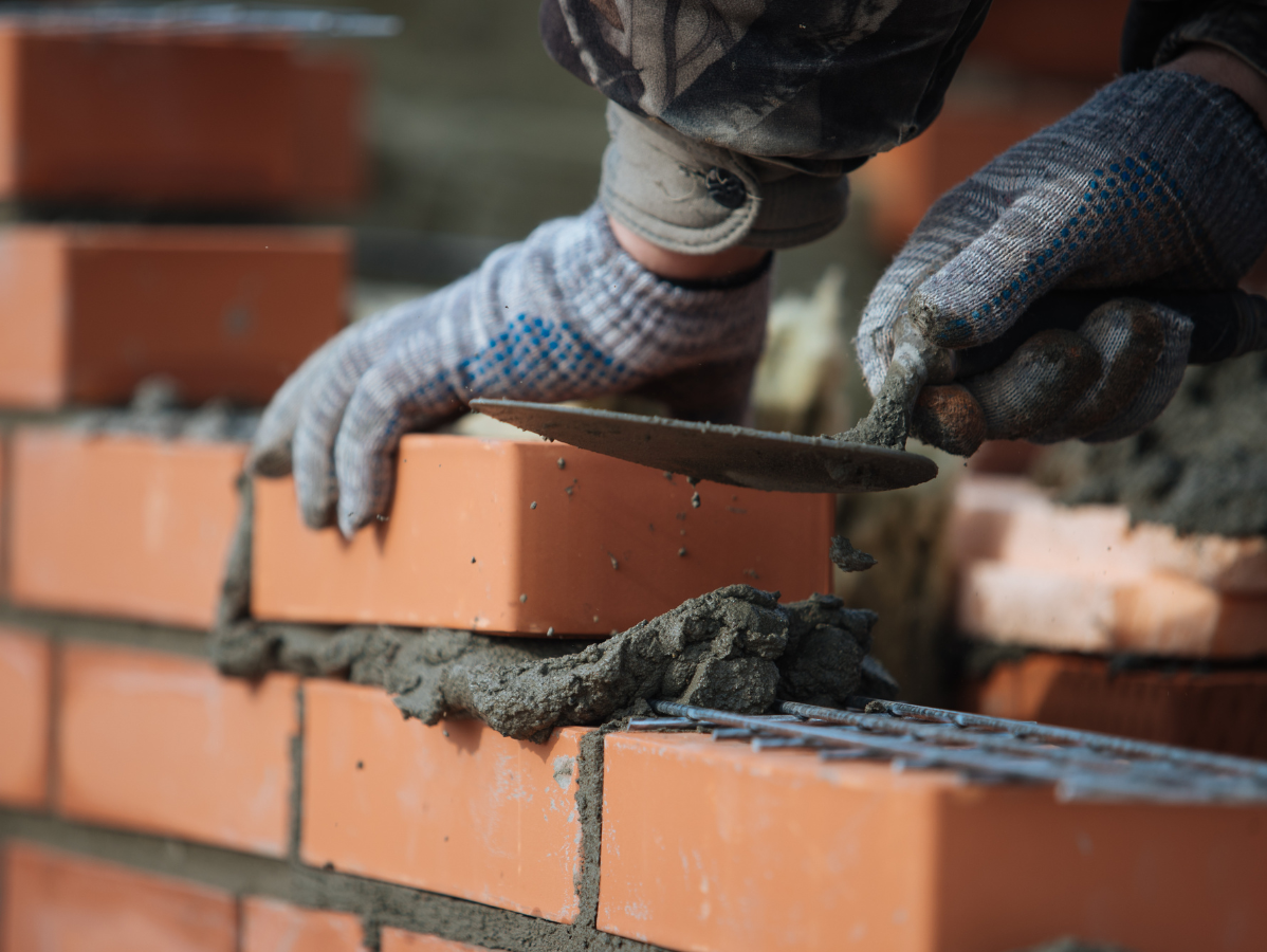 Gloved hands place a red brick onto a mortar-filled brick wall during construction