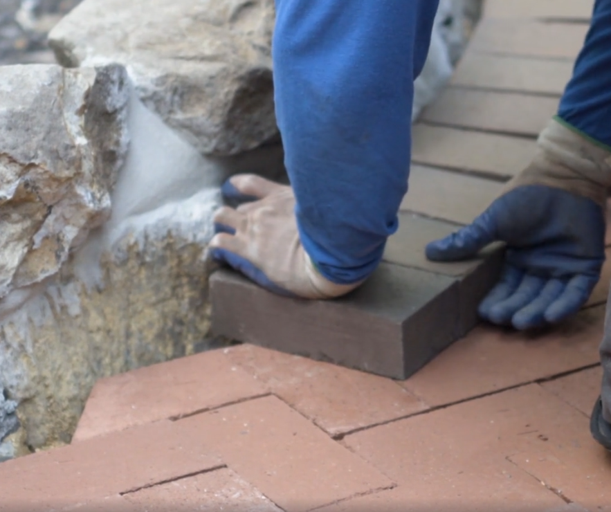 Hands installing a dark paver beside a stone wall on a brick walkway