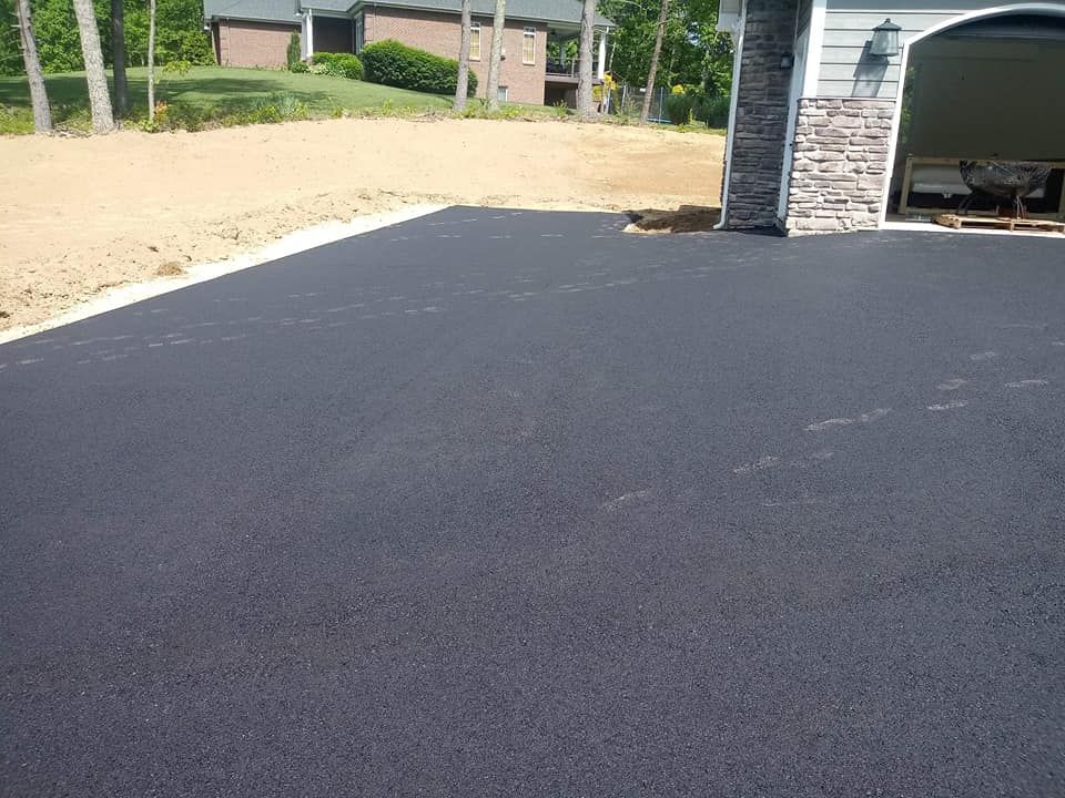 Newly paved black asphalt driveway next to a garage, with a house and bare dirt in the background.