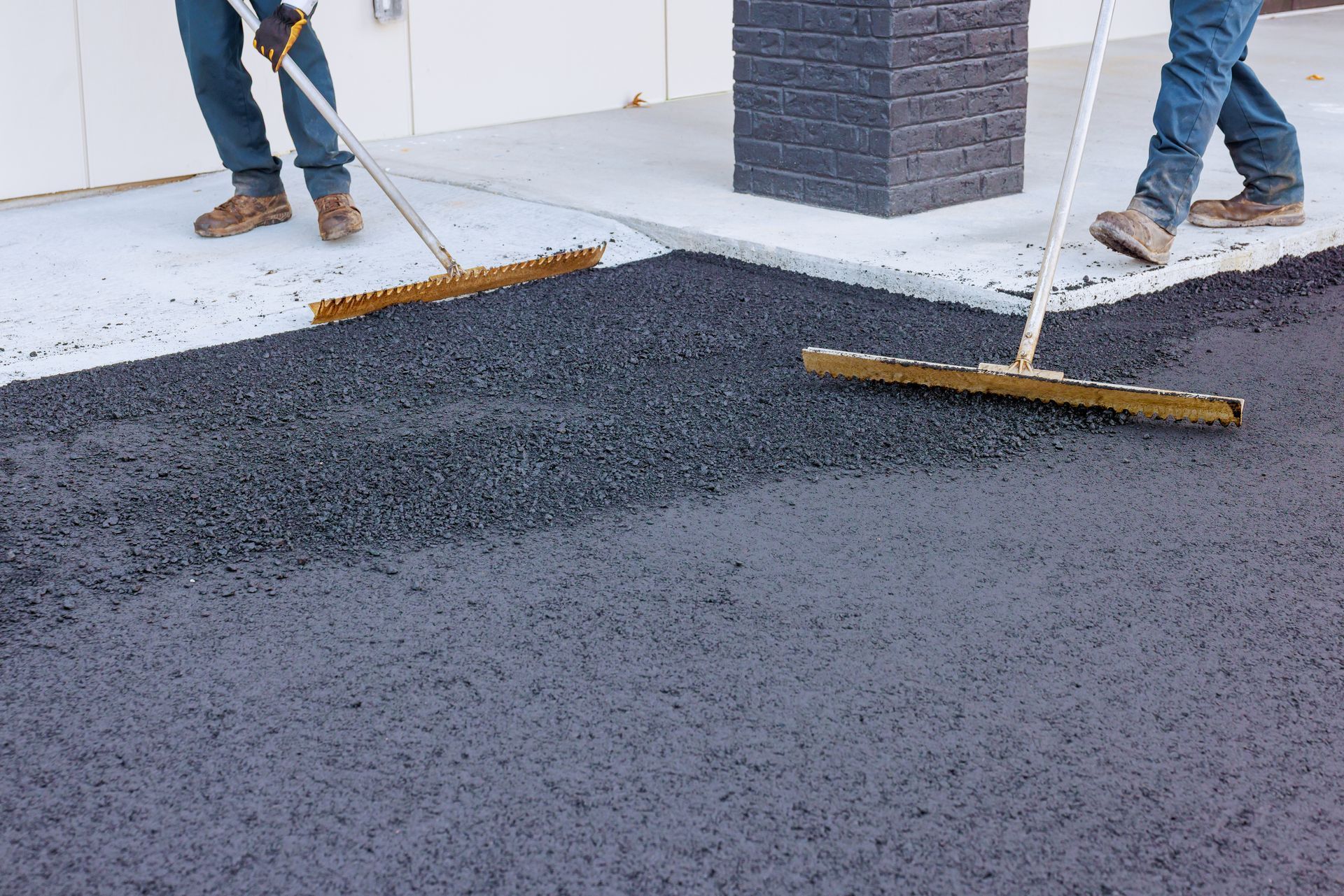 Workers smoothing freshly laid asphalt with rakes on a paved surface near a building.
