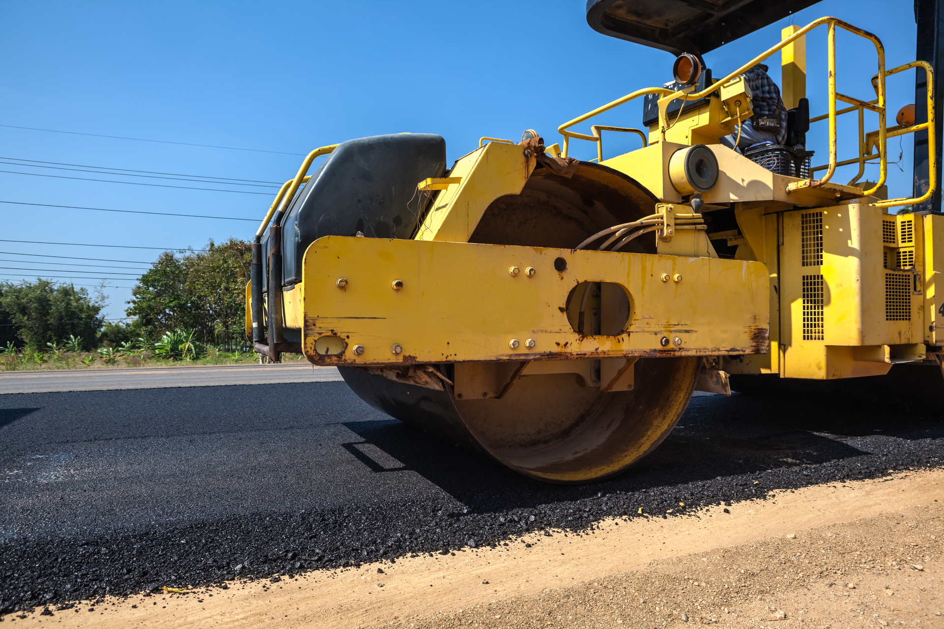 Yellow road roller compacting fresh asphalt during highway construction under clear blue sky.