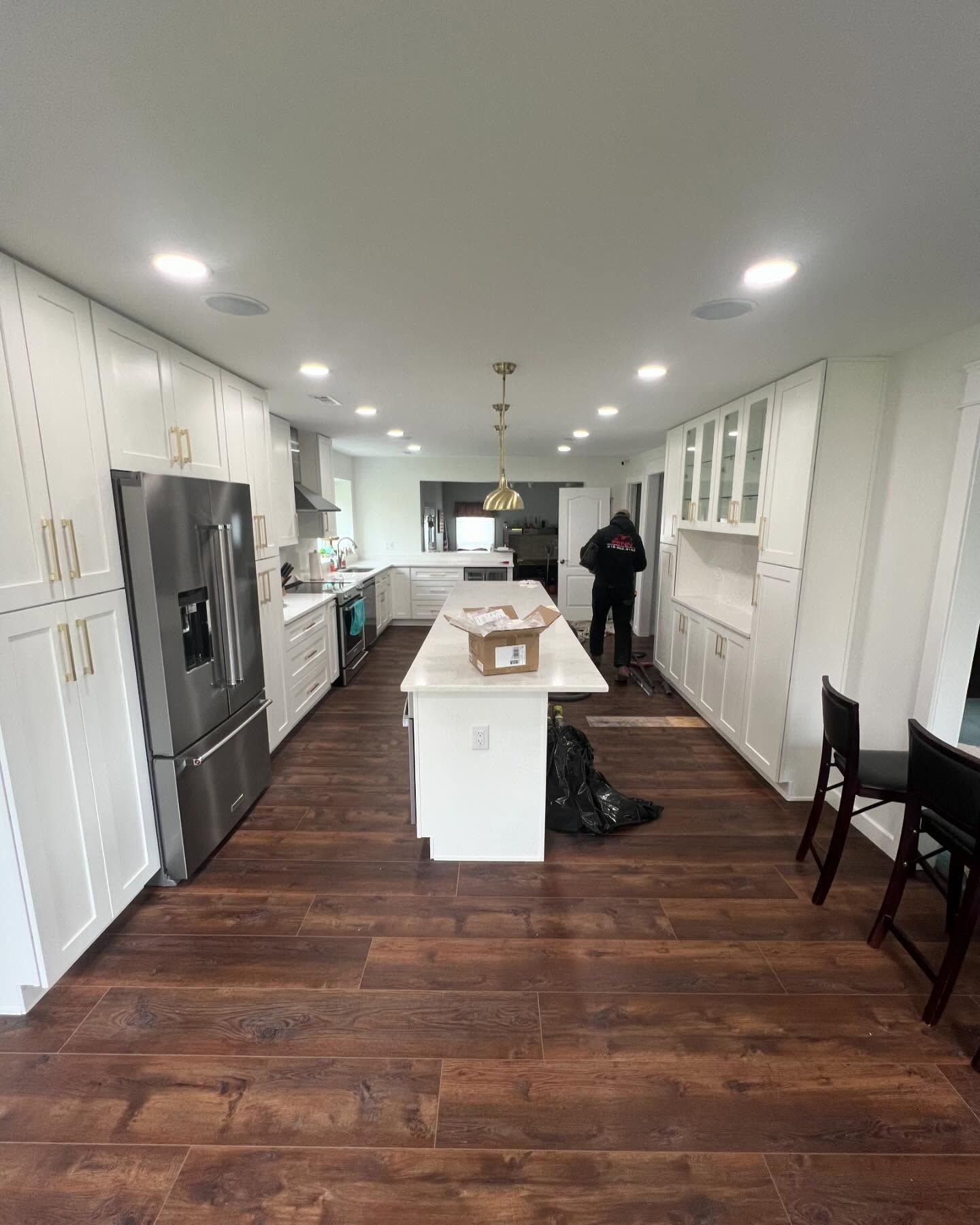 A renovated kitchen with white cabinets, dark wood floors, and a stainless steel refrigerator. A person works in the room.