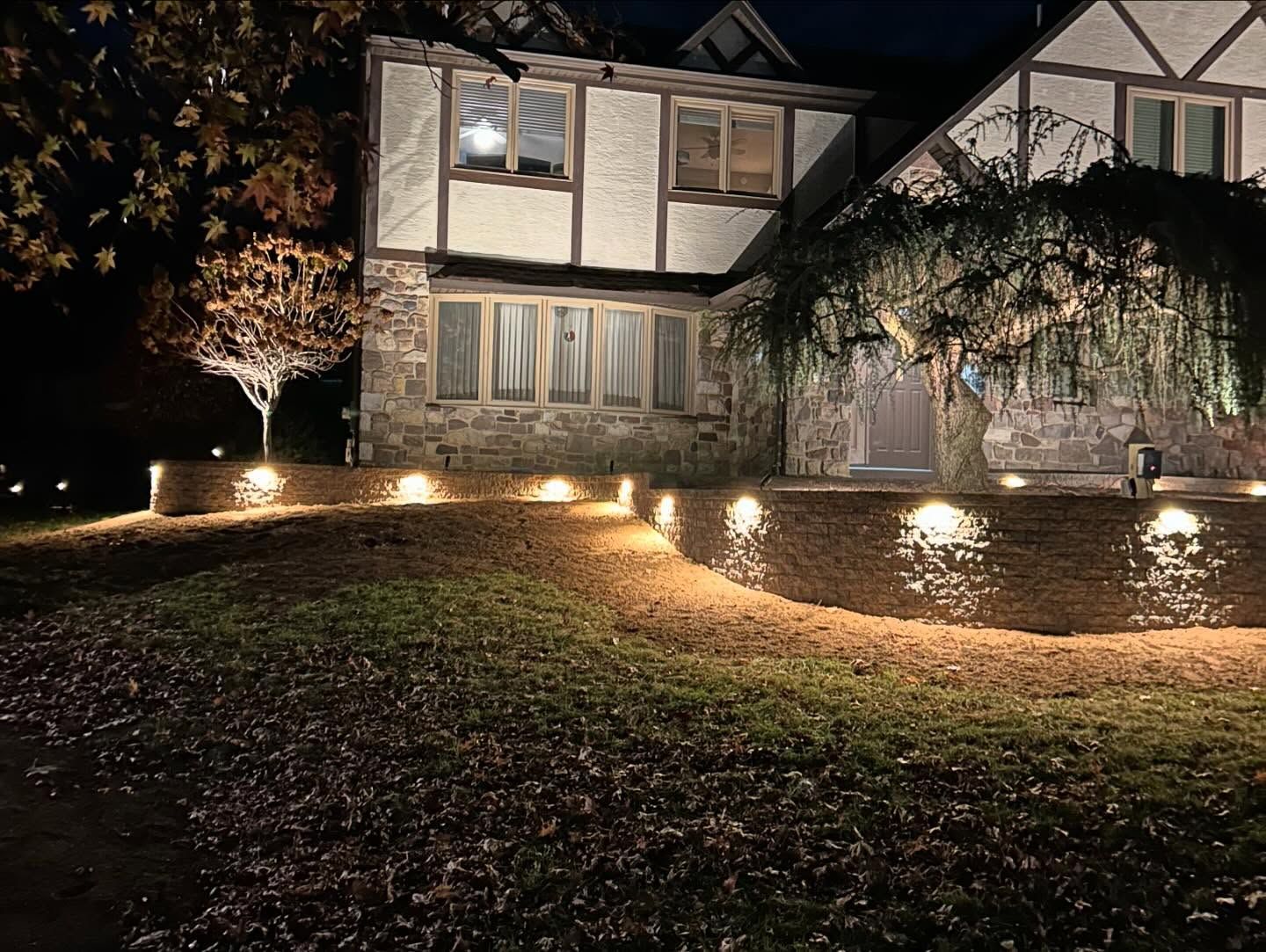 Tudor-style house lit at night with landscape lighting along a retaining wall and weeping tree.
