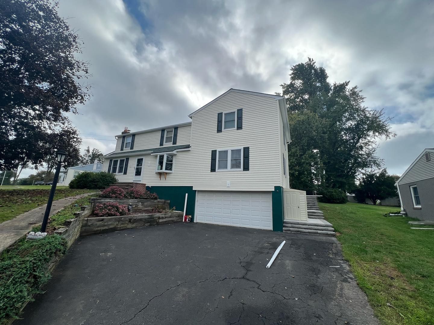 Two-story beige house with black shutters, white garage door, and black asphalt driveway under a cloudy sky.