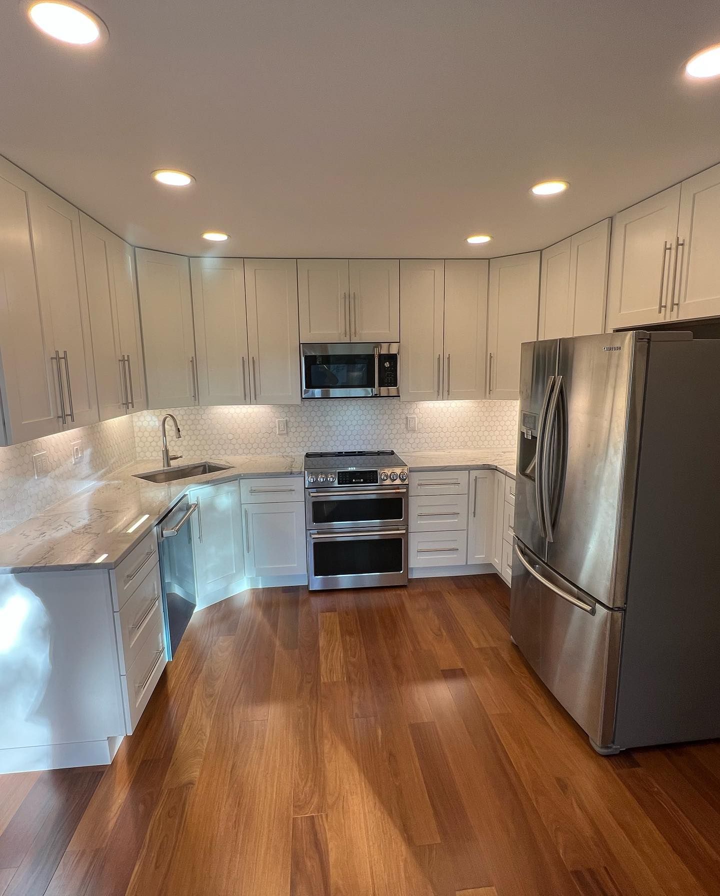 Modern white kitchen with stainless steel appliances, wood floor, and recessed lighting.