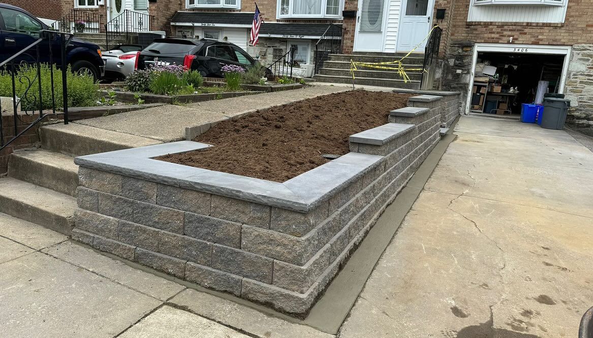 Retaining wall with soil, adjacent to a driveway and steps leading to a house.