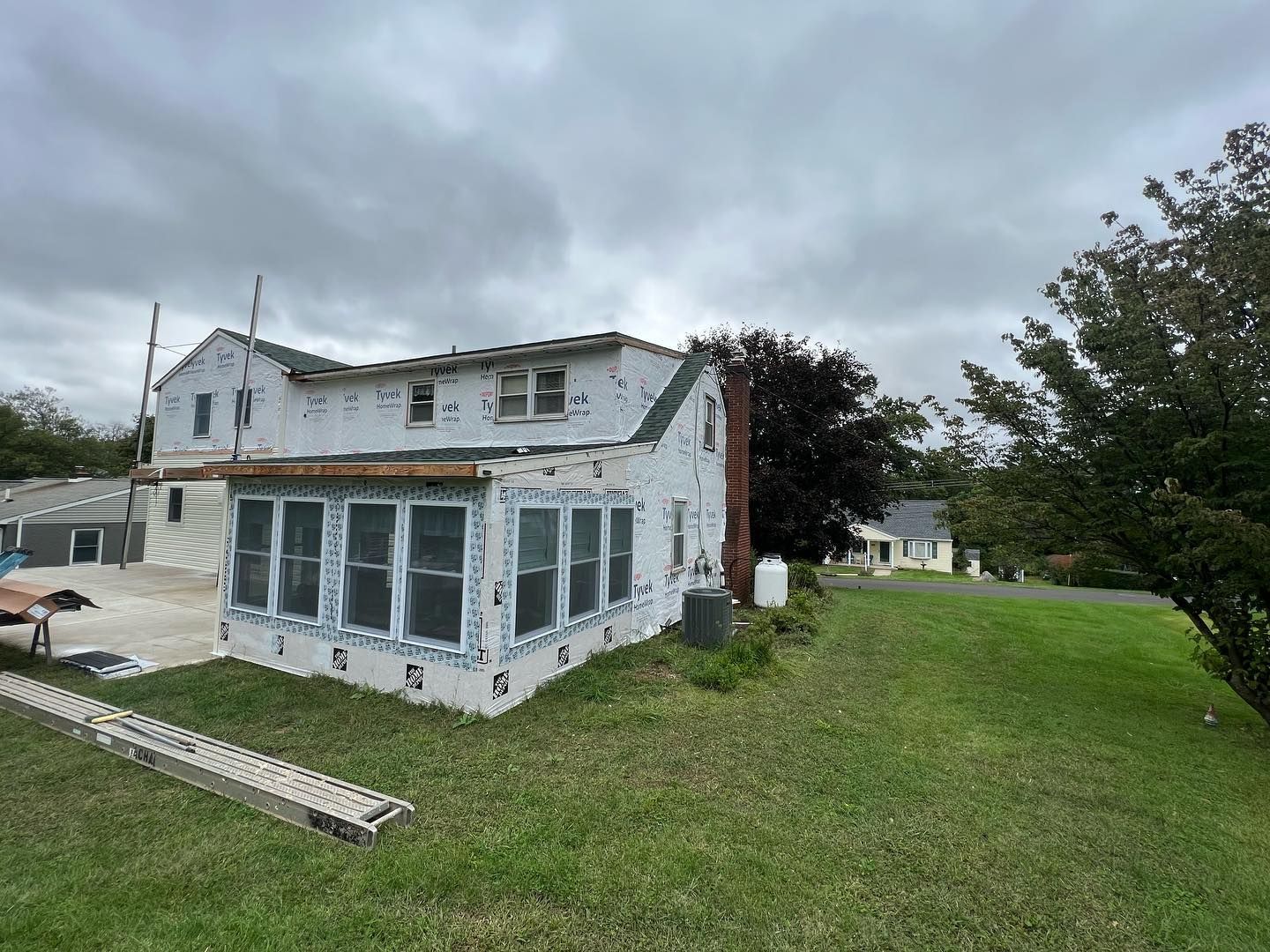 House with siding partially removed, revealing sheathing. Green grass, cloudy sky.