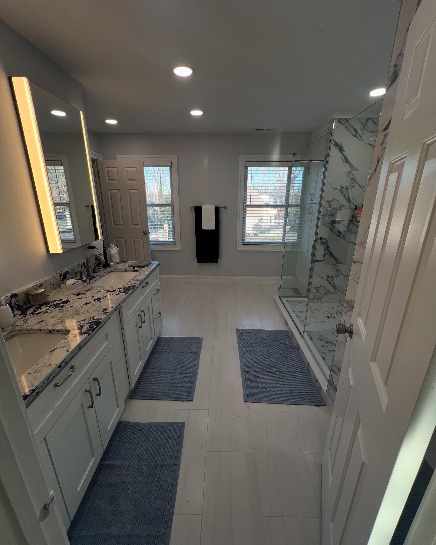 Bathroom with white cabinets, marble countertops, marble shower, and blue bath mats.