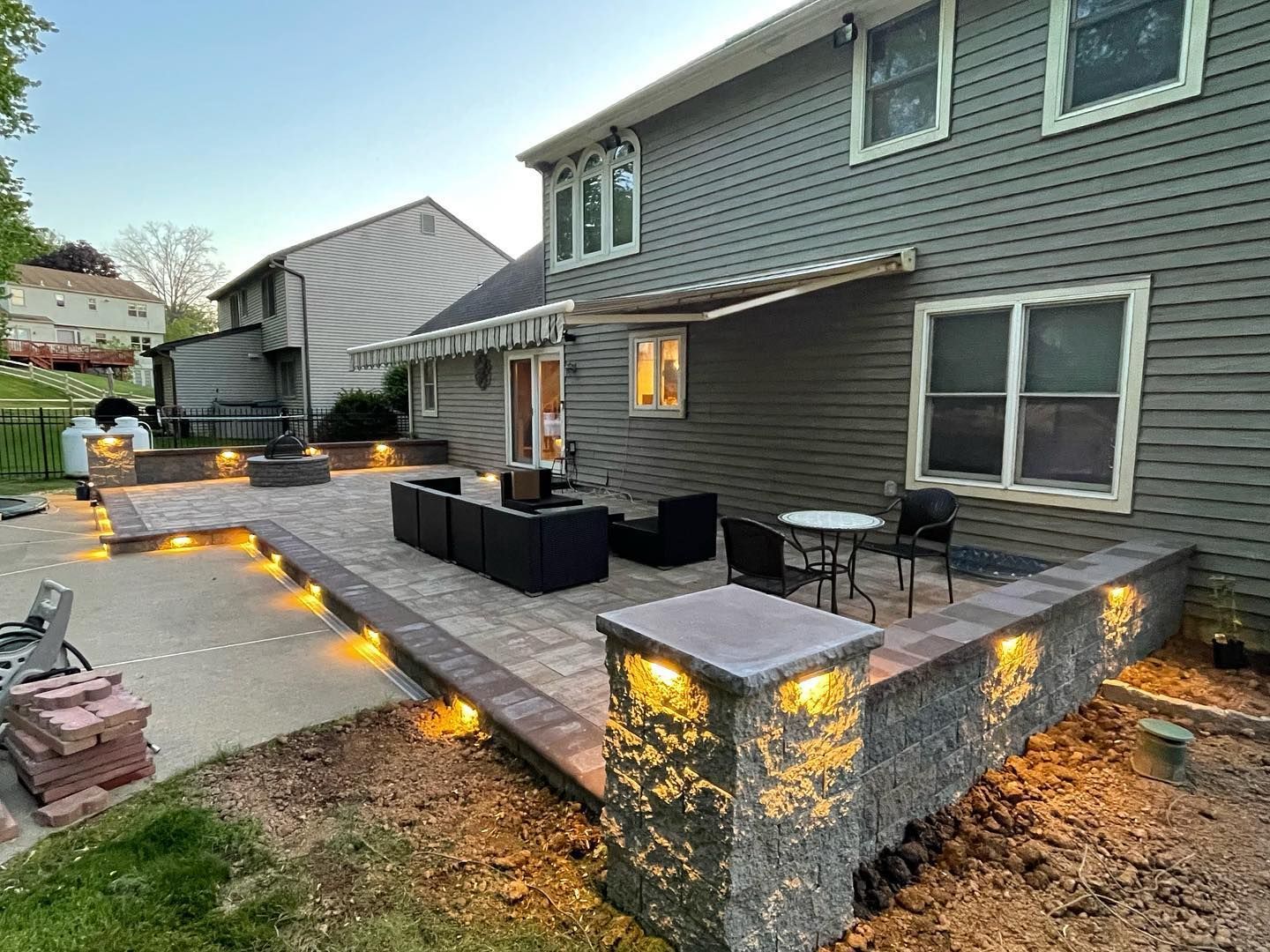 Backyard patio with retaining walls, seating areas, and lighting near a two-story house.
