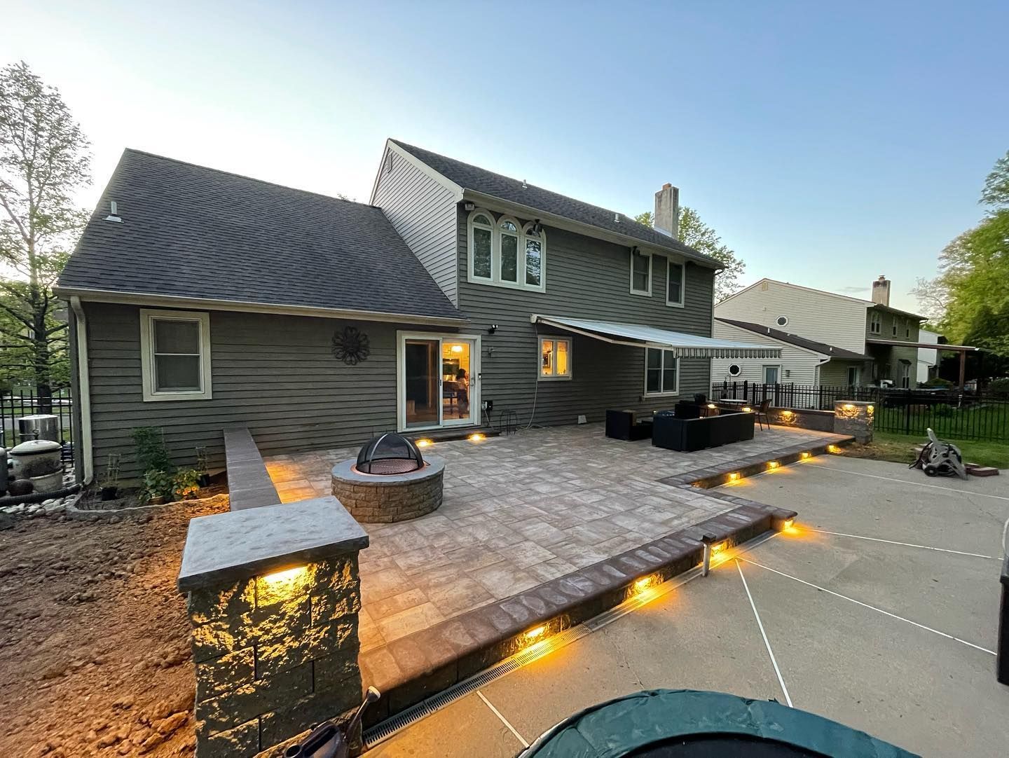 Backyard patio with fire pit, seating, and lighting next to a two-story house with gray siding.