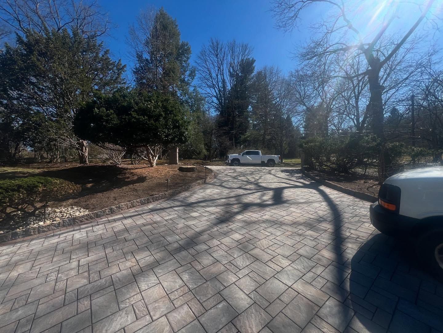 Driveway with brick pavers, trees, and two white vehicles parked. Bright sunlight.