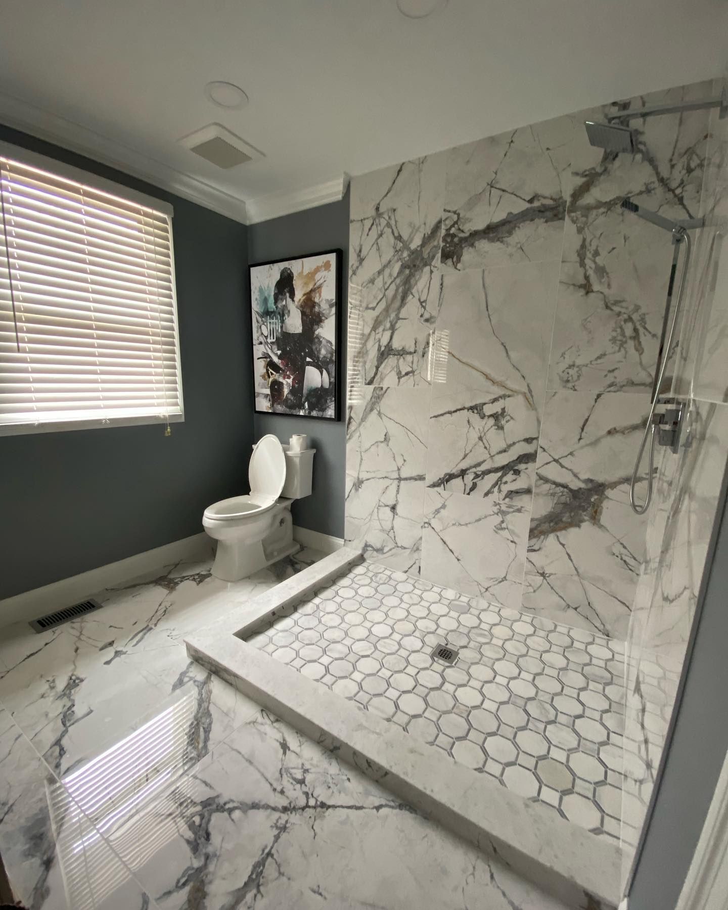 Modern bathroom with marble shower and flooring, toilet, art, and a window.