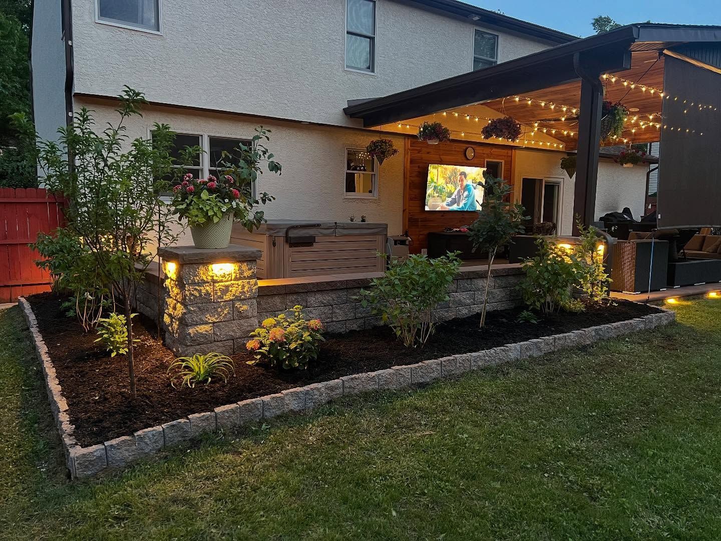 Backyard patio with a retaining wall, lights, flowers, and a TV mounted on a wooden structure.