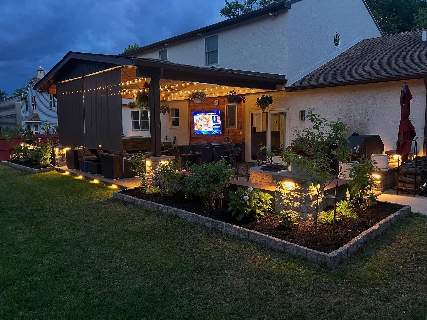 Backyard patio at night, with string lights, TV, and lit landscaping.