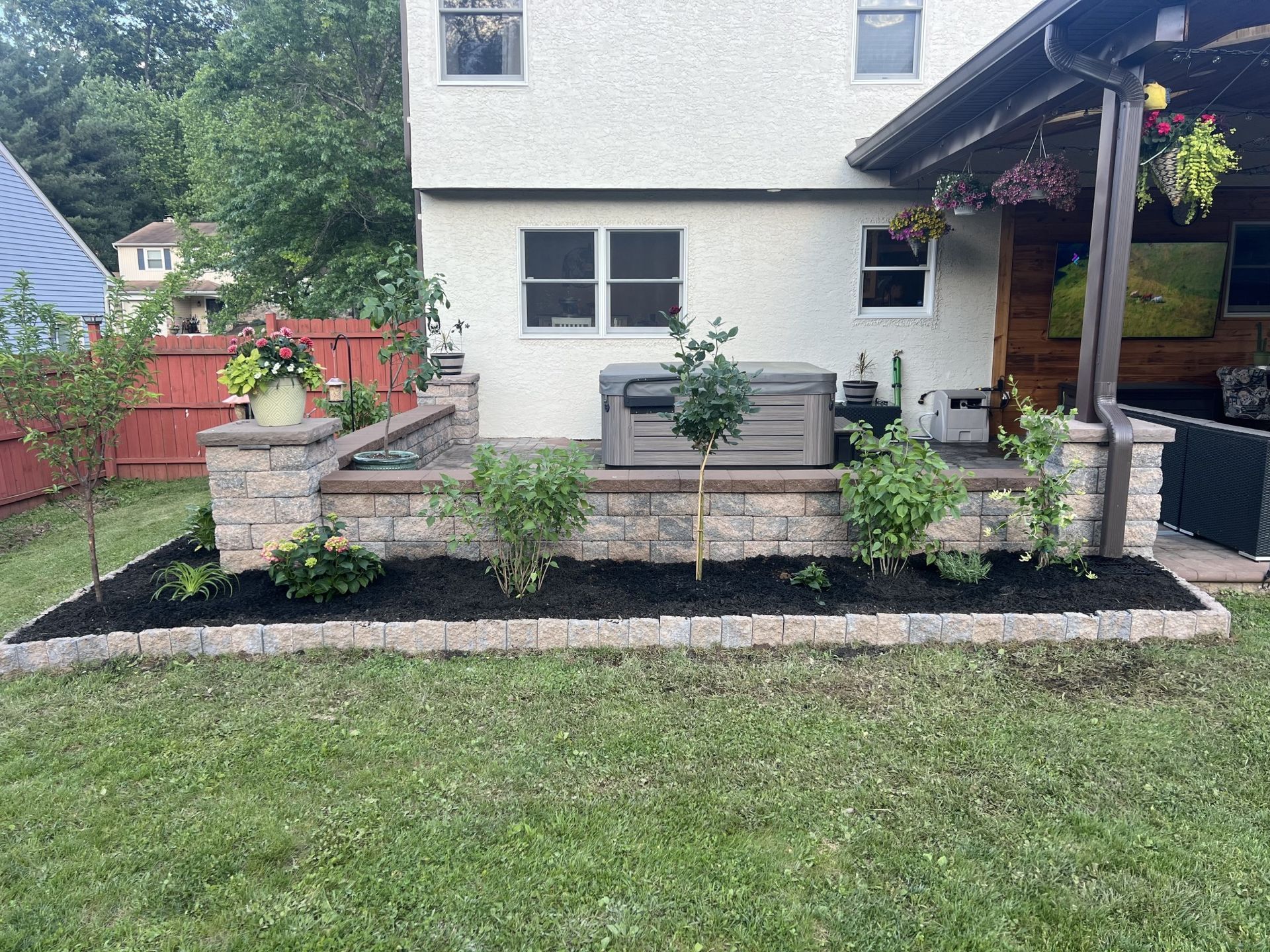 Backyard patio with retaining walls, a hot tub, and blooming plants against a two-story house.