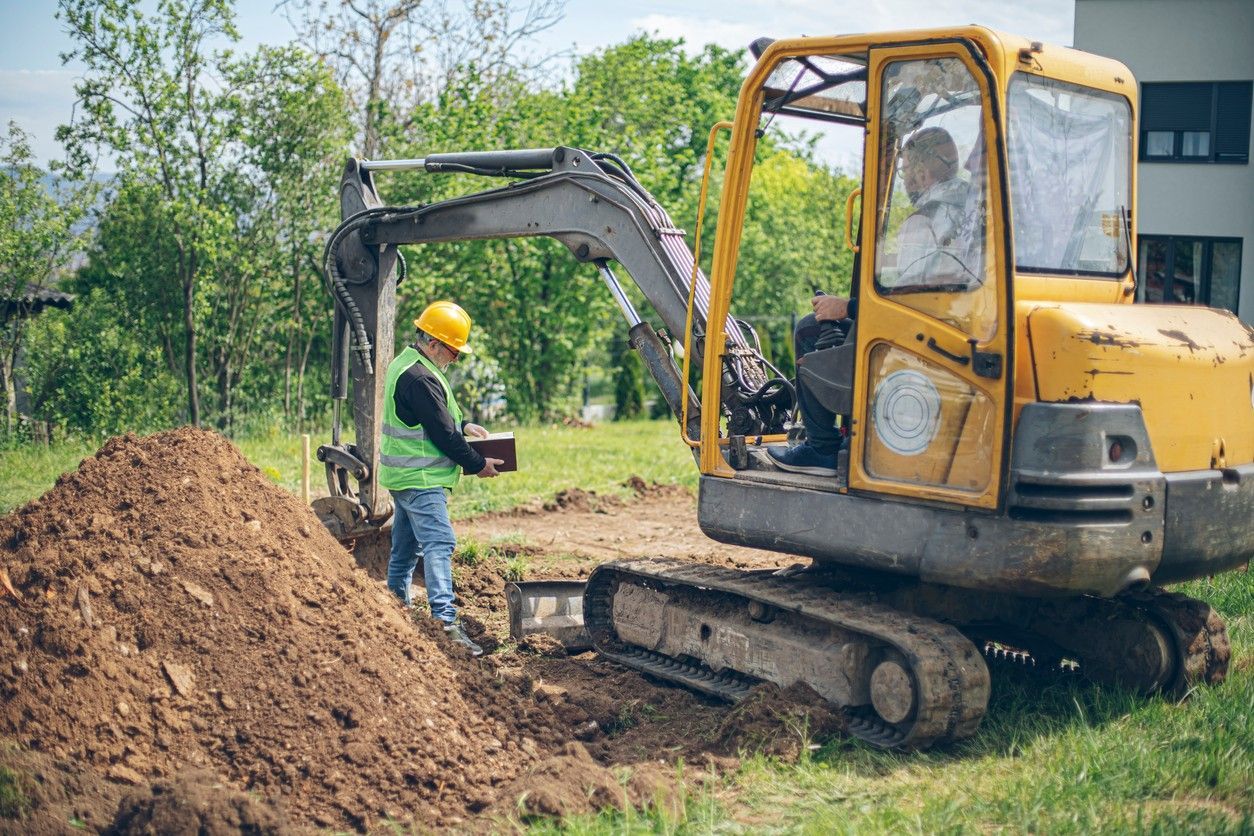 Construction worker at a job site with a yellow excavator, digging in the dirt.