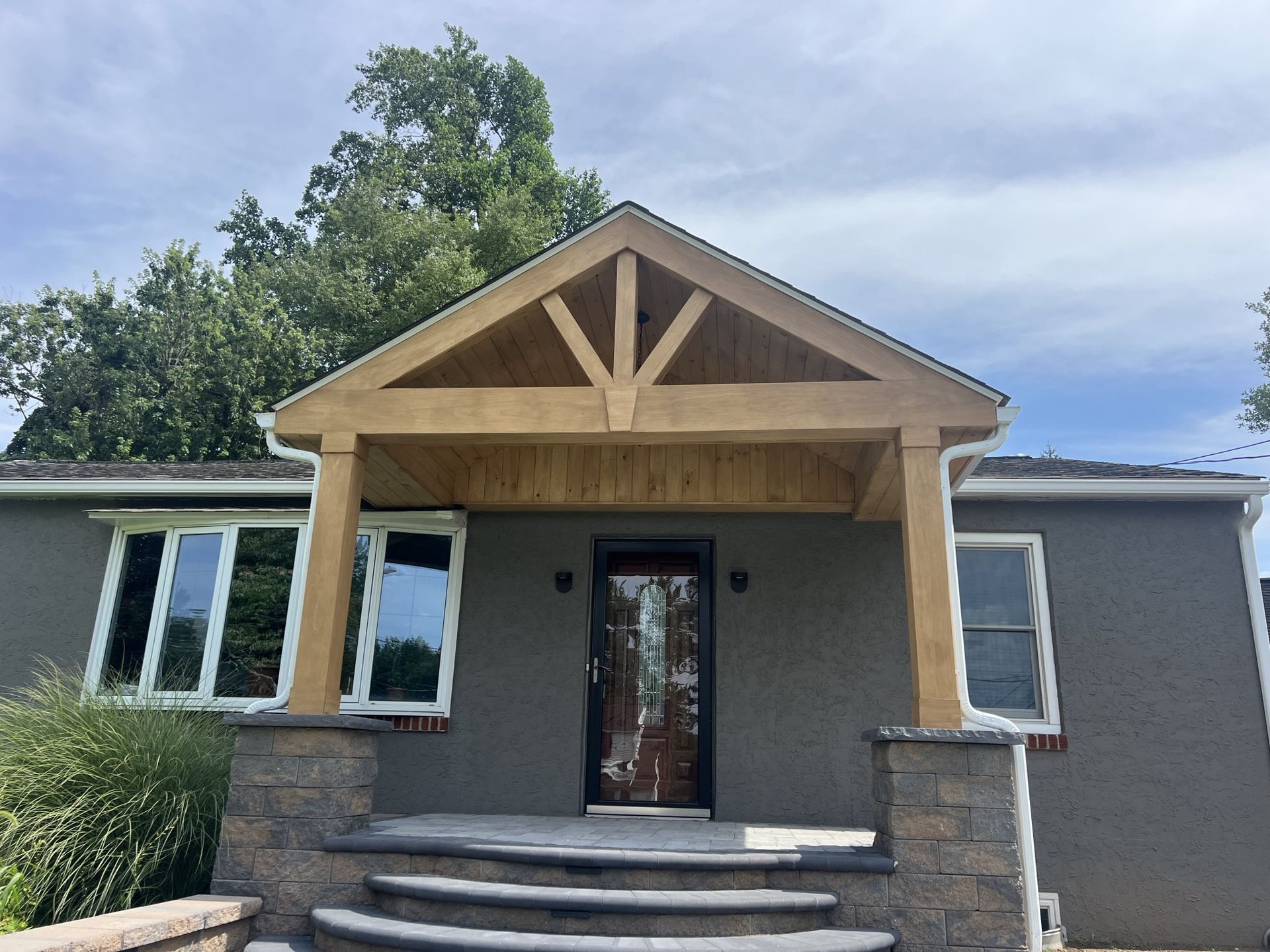 House entrance with a wooden porch, dark door, and stone pillars. Grey stucco exterior.