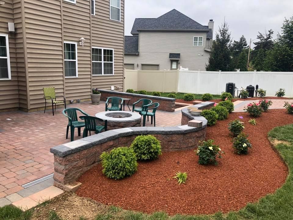 Backyard patio with fire pit, seating, retaining wall, and landscaped flower beds with red mulch.