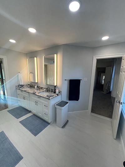 Bathroom with double sink vanity, gray walls, white floor, blue rugs, and door.