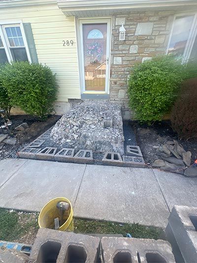 Construction of a stone walkway leading to a house entrance. Cinder blocks, stone, and a yellow bucket are visible.