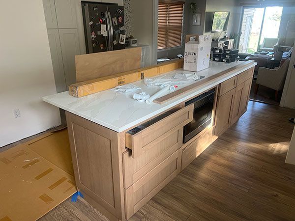 Kitchen island with white countertop, light wood cabinets, and built-in microwave, under construction.