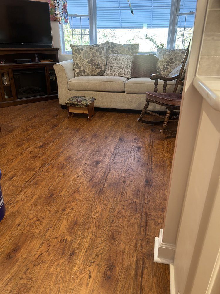Wooden floor in a living room with sofa, ottoman, and rocking chair near a window.