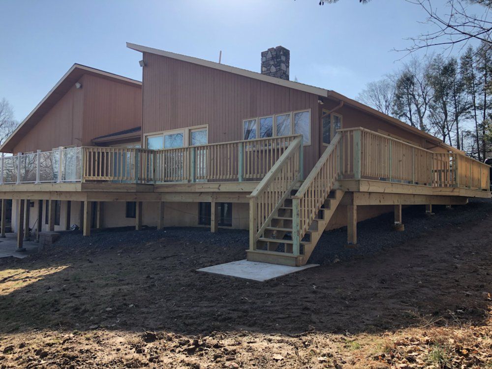 Wooden deck wraps around a brown house with glass and wood railings, stairs, and a concrete pad on a hillside.