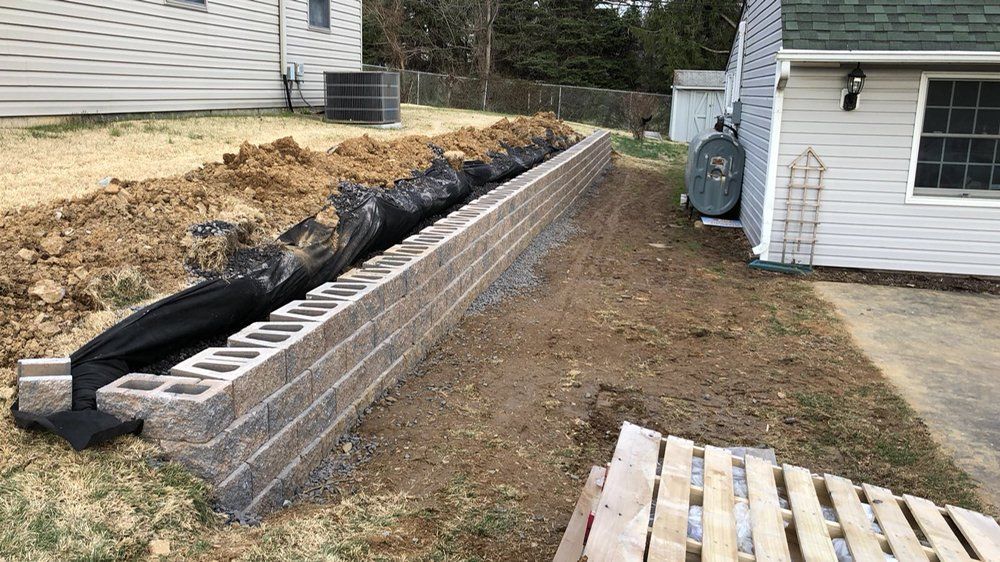 A retaining wall made of stacked concrete blocks under construction, next to a house. Soil and black fabric are visible.
