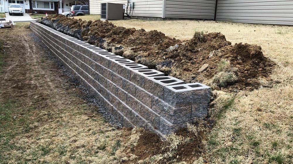 Retaining wall made of gray concrete blocks under construction, with dirt pile and dry grass.