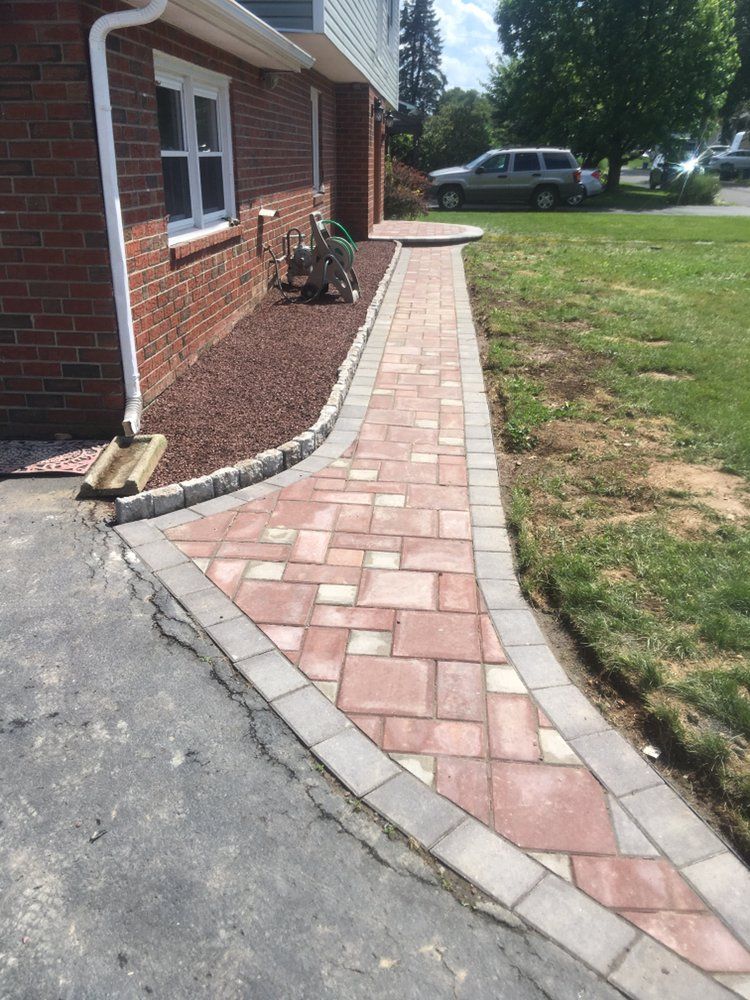 Brick pathway bordered by gray pavers beside a brick house and grassy yard.