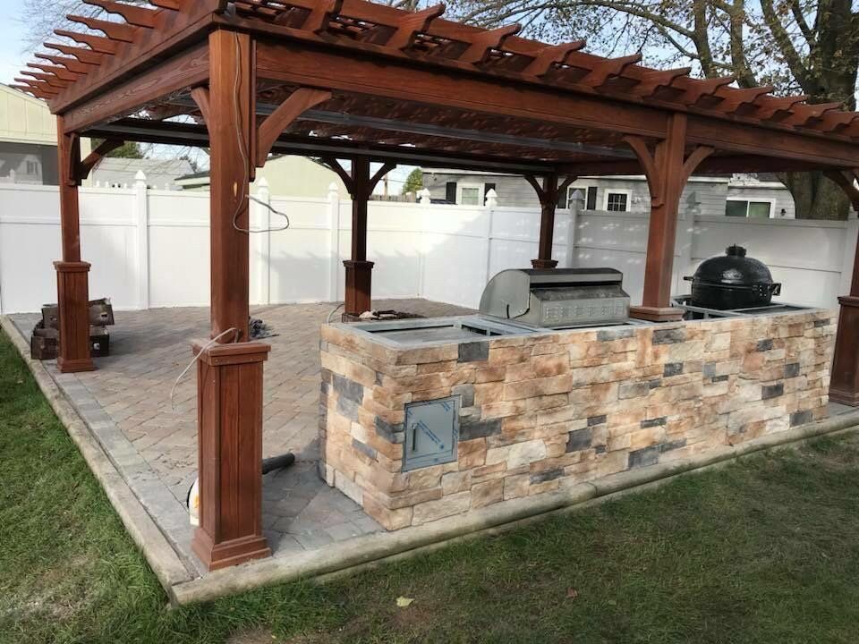 Outdoor kitchen with stone facade, under a wooden pergola, housing a grill and a smoker.